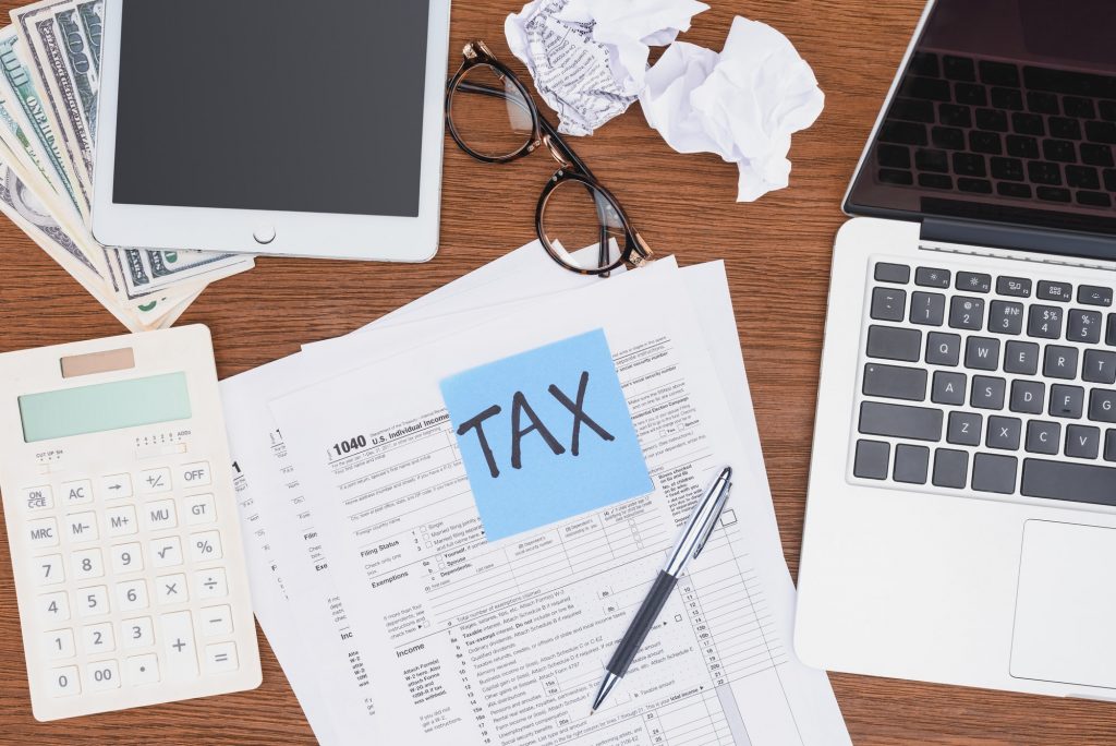 top view of tax forms, digital devices and blue card with 'tax' word on desk